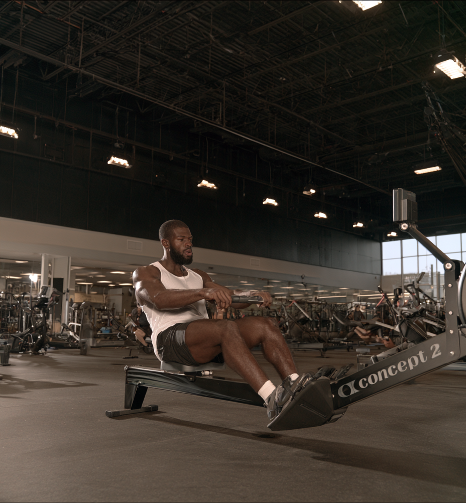 A man in a white tank top rowing on a Concept2 rowing machine in a large, open gym