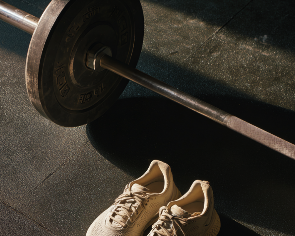 Overhead view of a loaded barbell and a pair of cream sneakers on a dark rubber gym floor in warm light